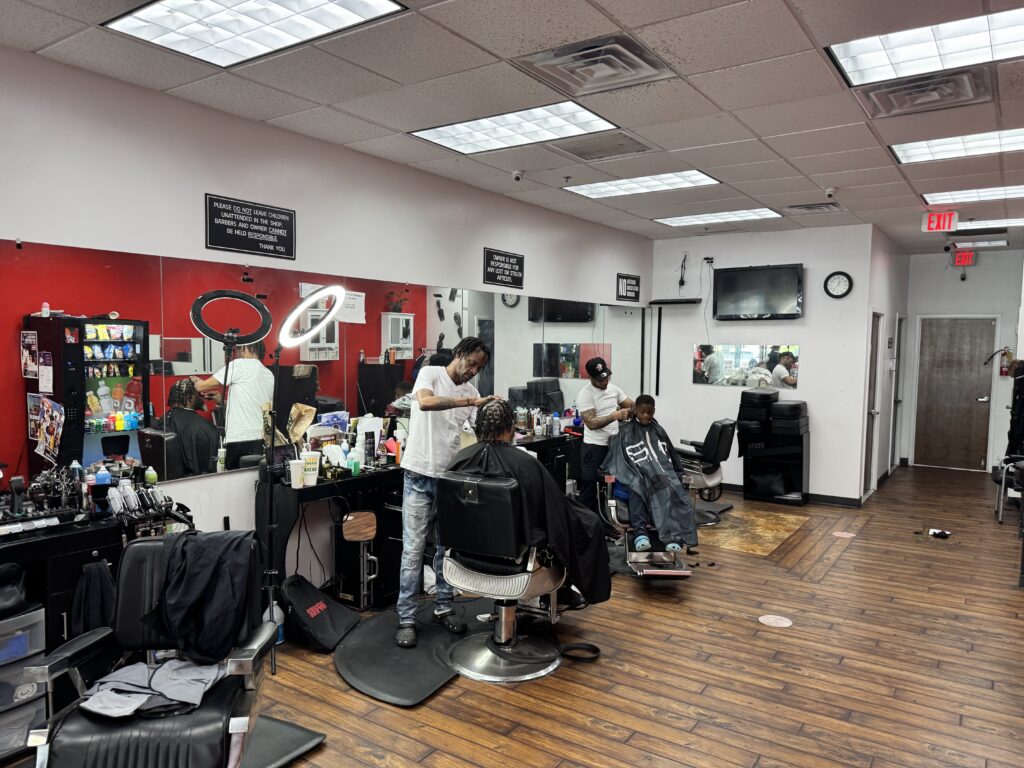 Two barbers cut hair for clients in a modern barbershop at the Shops at Park Village, featuring red and white walls, large mirrors, wooden floors, and various barber tools on the counters.