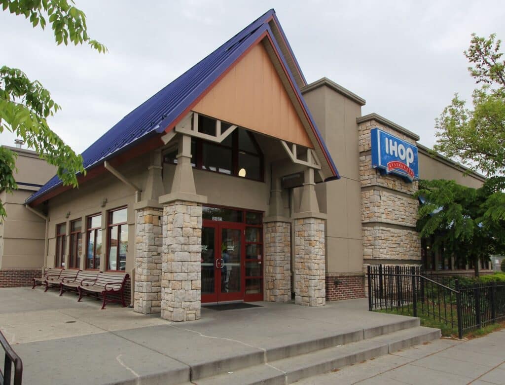 Exterior of an IHOP restaurant with stone pillars, large blue sign, and a sloped roof. Entrance has glass doors and adjacent benches.