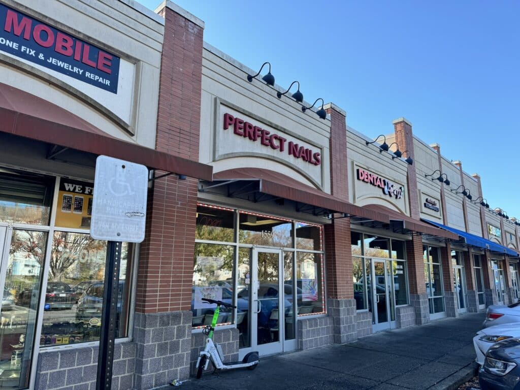 A row of businesses including a mobile repair shop, nail salon, and dental office on a sunny day. A scooter is parked on the sidewalk.