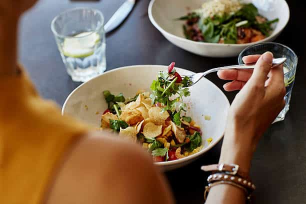 A person eating a salad with potato chips and greens at a table, with a glass of water nearby and another bowl of food in the background.