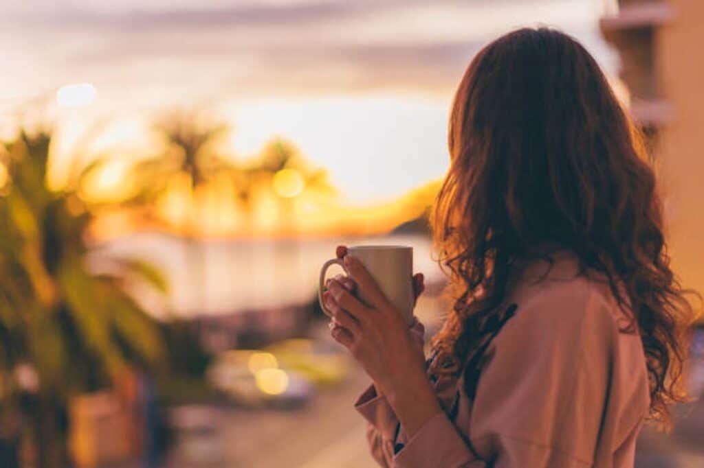 A person holding a mug looks at a sunset over a coastal landscape with palm trees.