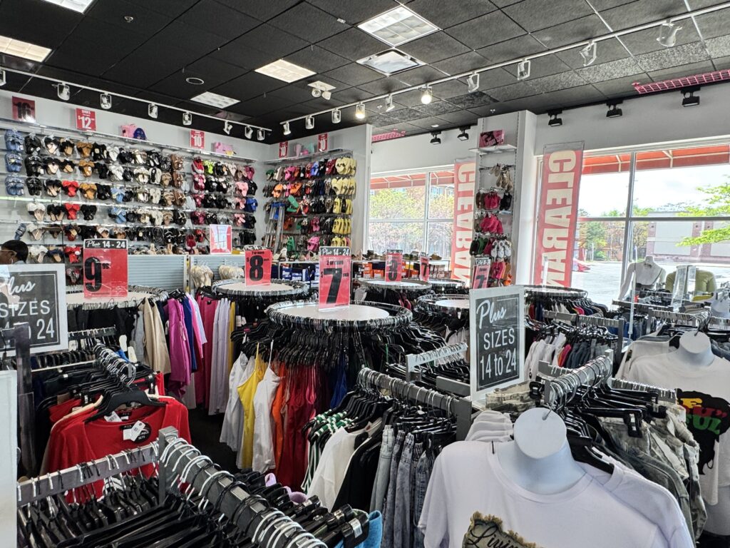 Clothing store interior with racks of assorted women’s clothes, mannequins in the foreground, and walls displaying wigs and hats. Signs show sizes and price sections—perfect for finding a thoughtful Mother's Day gift.