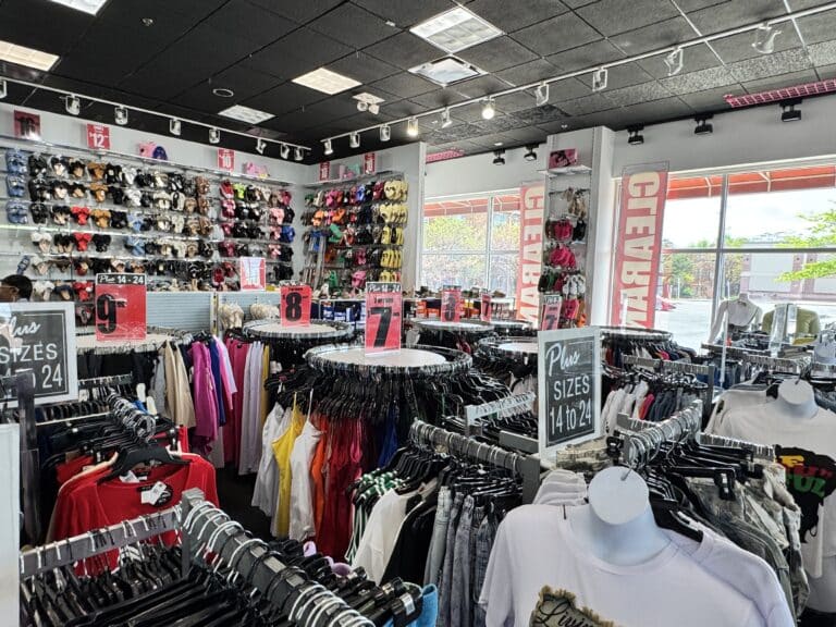 Clothing store interior with racks of assorted women’s clothes, mannequins in the foreground, and walls displaying wigs and hats. Signs show sizes and price sections—perfect for finding a thoughtful Mother's Day gift.