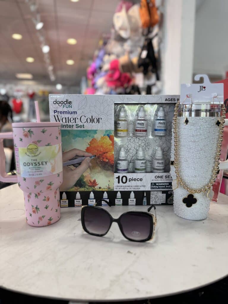 A set of three tumblers, a box of watercolor paints, and a pair of black sunglasses displayed on a round white table in a store.