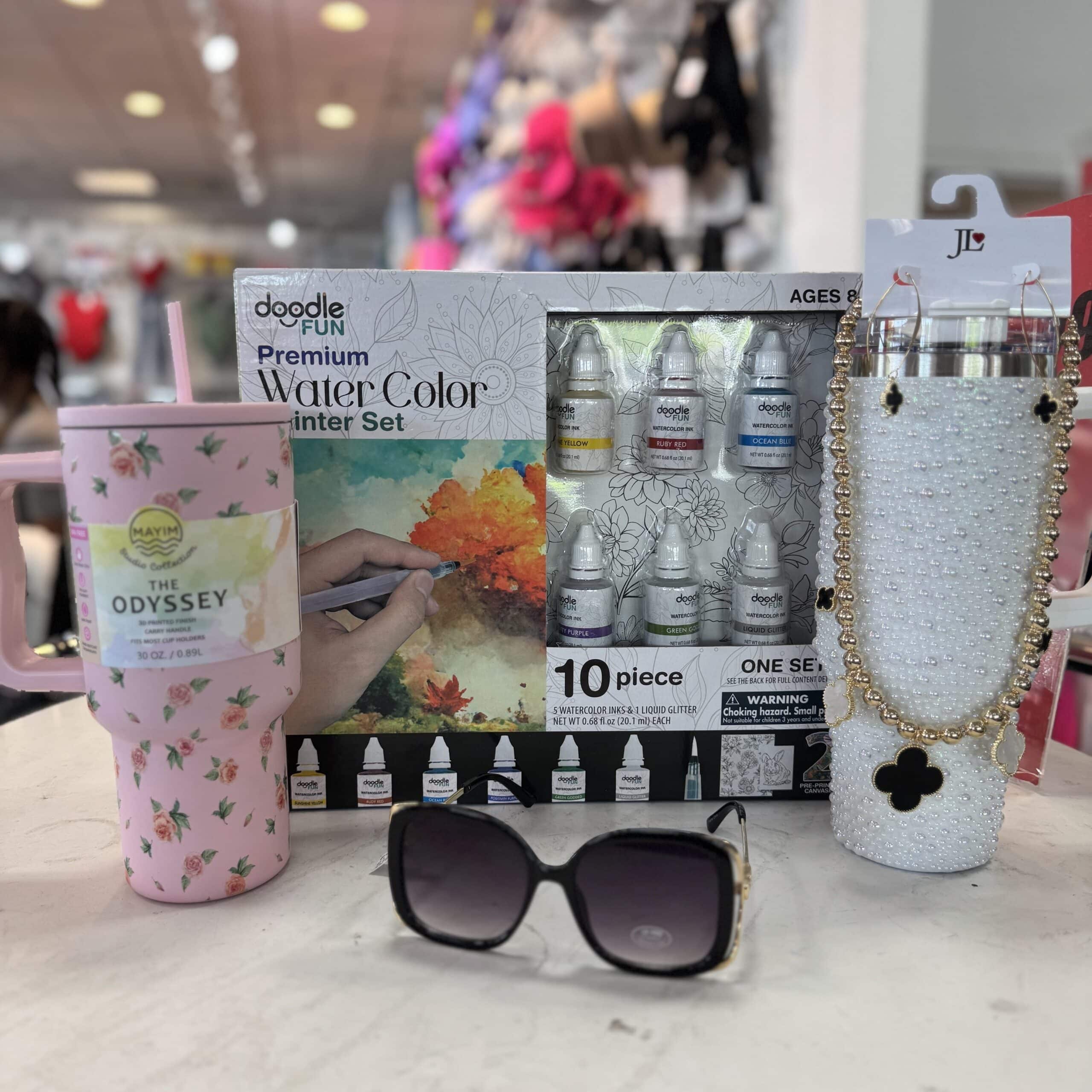 A floral tumbler with a book, a box of watercolor paints, black sunglasses, and a rhinestone tumbler with a cross charm displayed on a white table in a store.